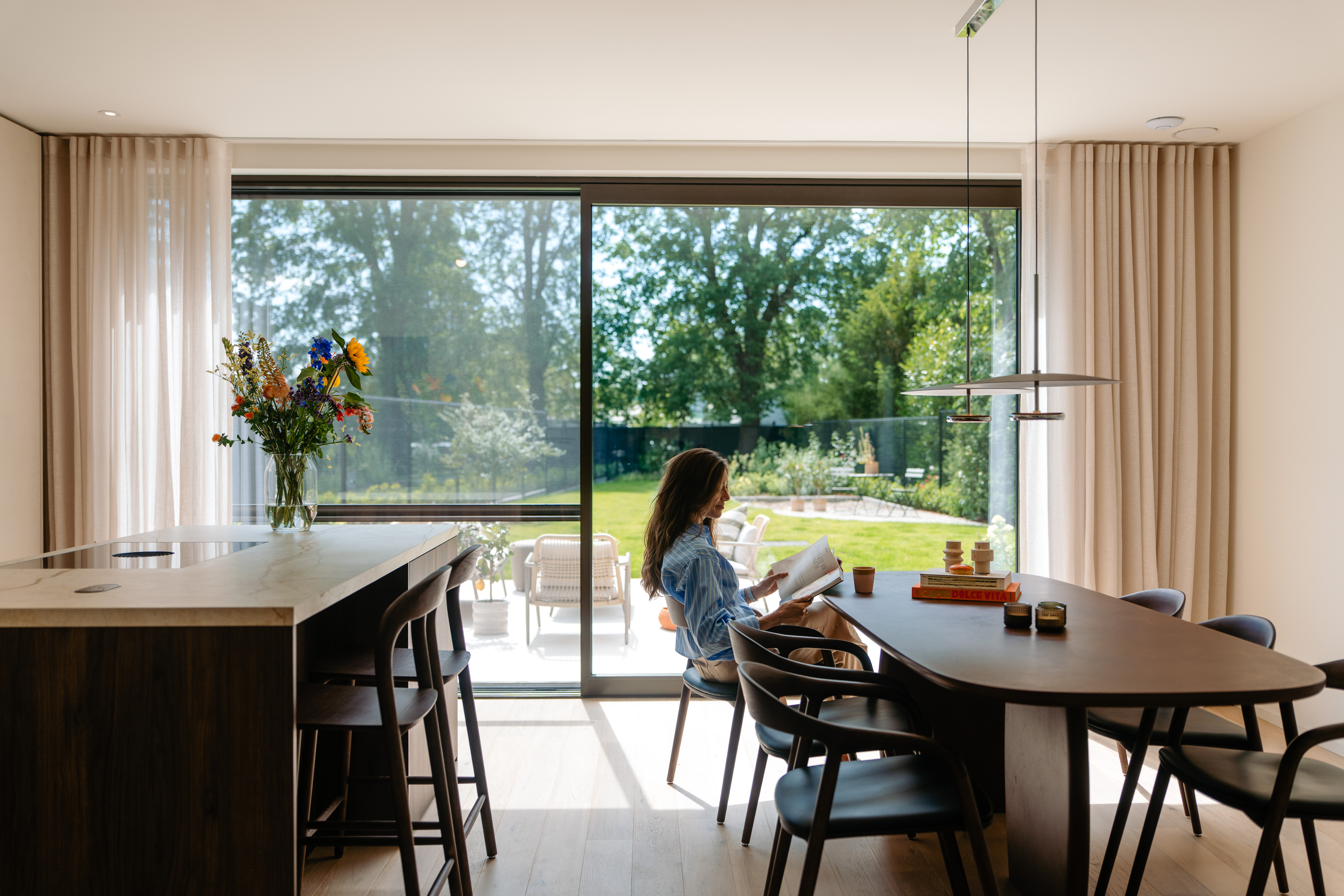 De afbeelding toont een moderne eetruimte met grote schuiframen die uitkijken op een weelderige tuin. De ramen laten veel natuurlijk licht binnen, wat zorgt voor een heldere en luchtige sfeer. Het strakke ontwerp van de schuiframen past perfect bij het eigentijdse interieur met een houten eettafel en stoelen.