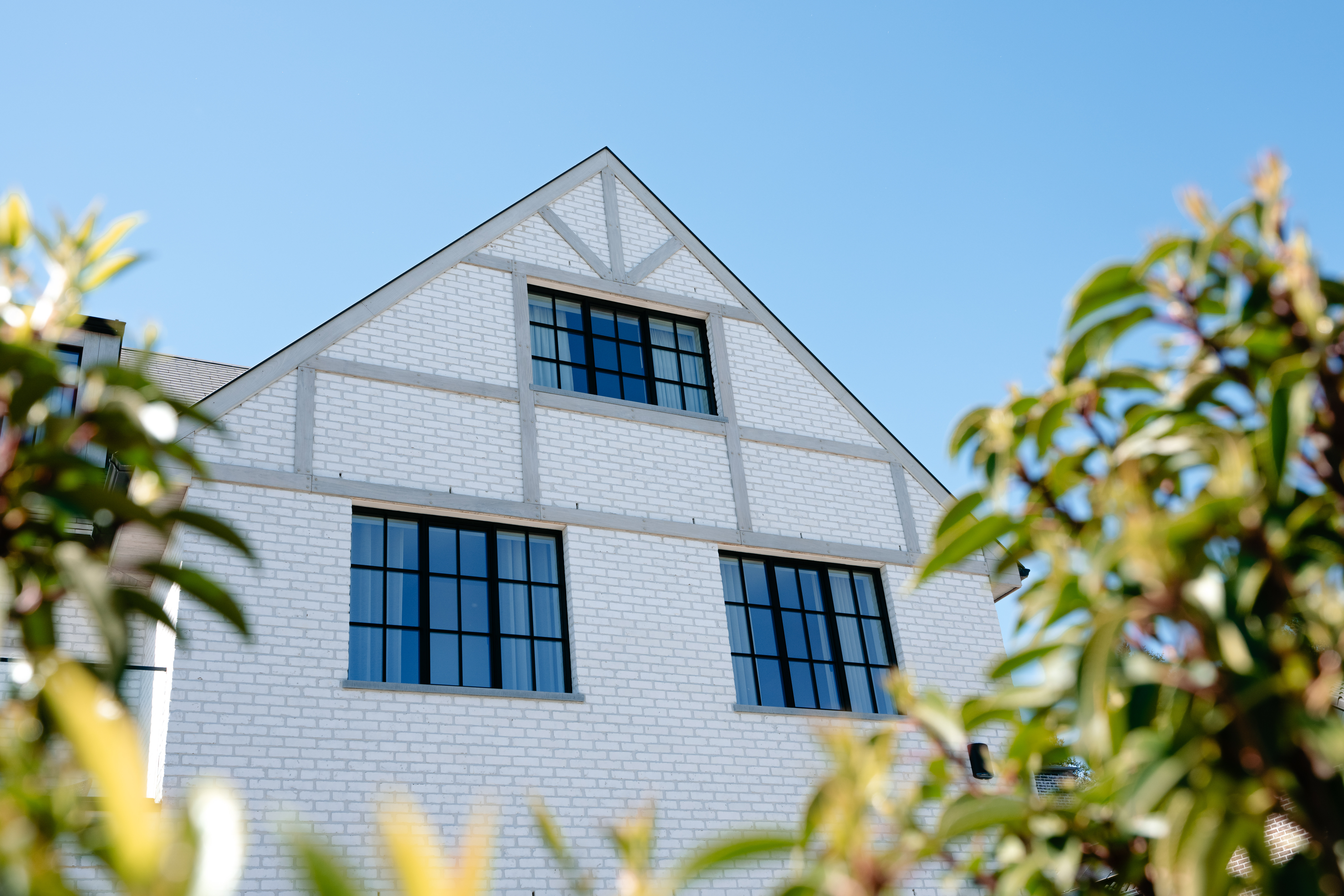De afbeelding toont een modern huis met een witte bakstenen gevel en grote, zwartomrande ramen. De ramen zijn symmetrisch geplaatst, wat het architecturale ontwerp versterkt. De heldere blauwe lucht vormt een opvallend contrast met de witte bakstenen en donkere raamkozijnen.