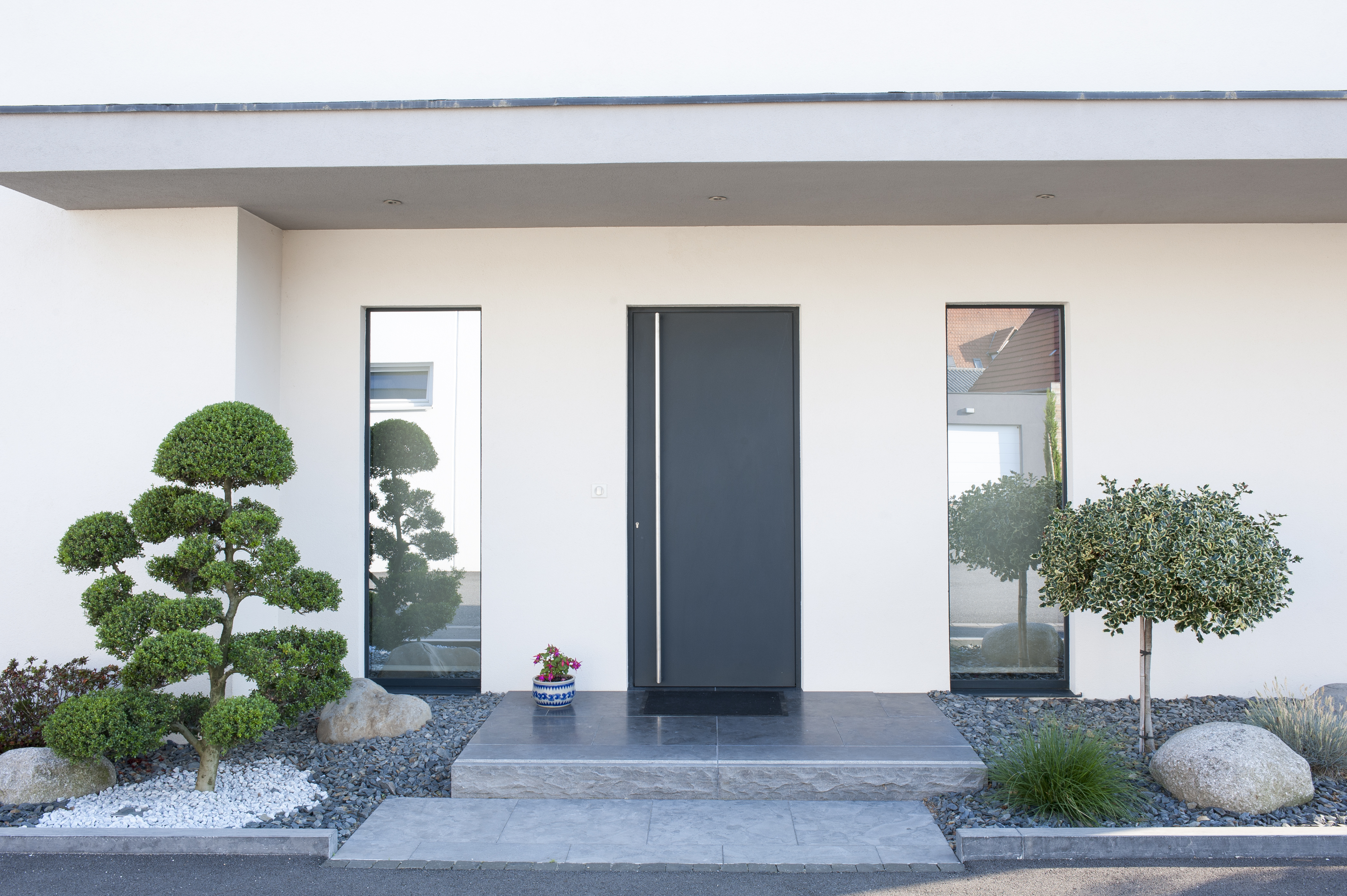 De afbeelding toont een moderne ingang met een strakke zwarte deur, geflankeerd door twee hoge, smalle ramen. De ramen zijn reflecterend, wat diepte en elegantie aan de gevel toevoegt. Het minimalistische ontwerp wordt aangevuld door netjes gesnoeide struiken en een schoon stenen pad dat naar de ingang leidt.