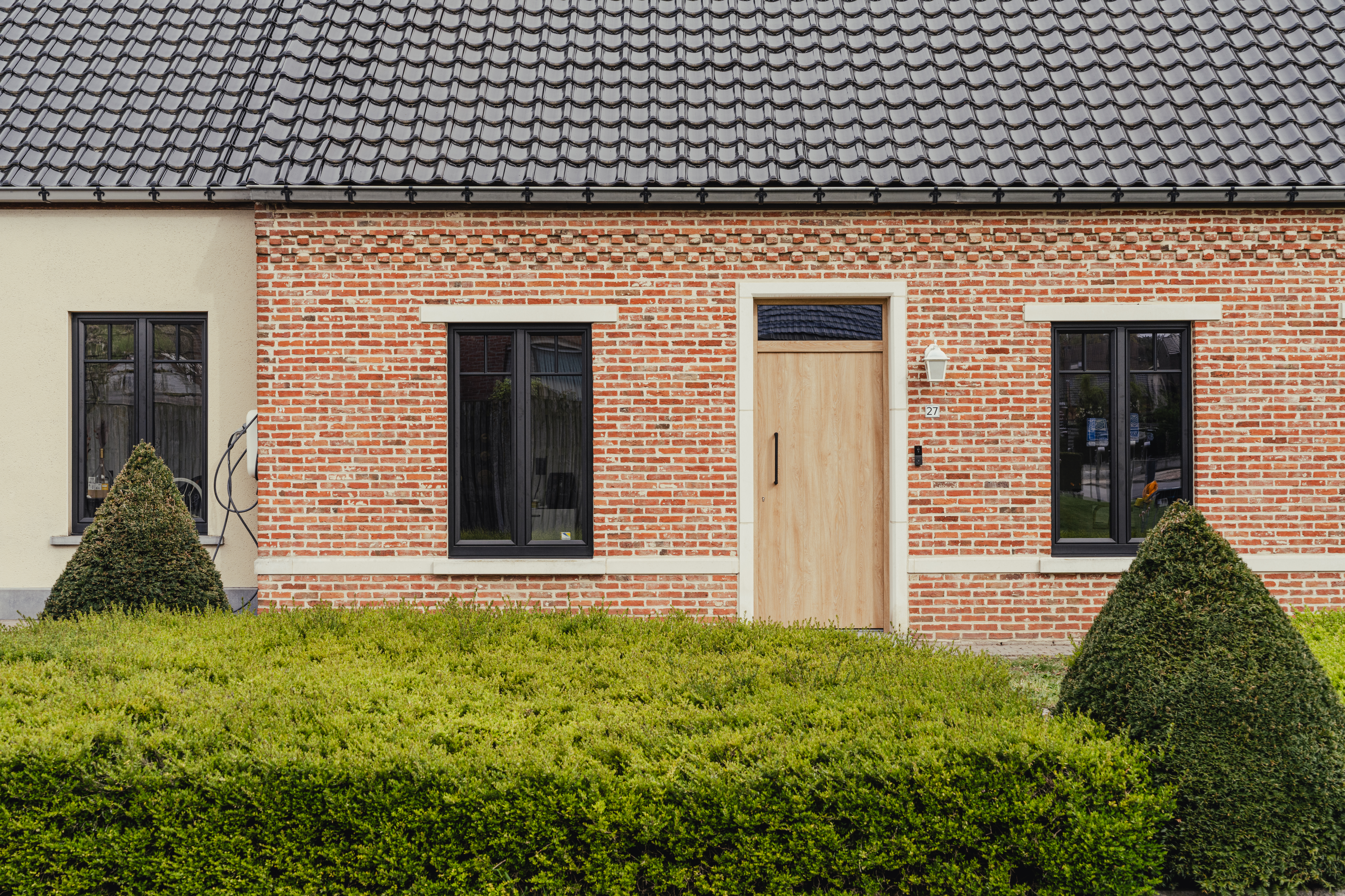 De afbeelding toont een charmant bakstenen huis met een modern ontwerp. De gevel heeft drie zwartomrande ramen en een houten deur, wat een evenwichtige en uitnodigende uitstraling geeft. De ramen zijn symmetrisch geplaatst, wat de esthetische aantrekkingskracht vergroot. De houten deur vult het metselwerk aan en voegt warmte toe aan het geheel.