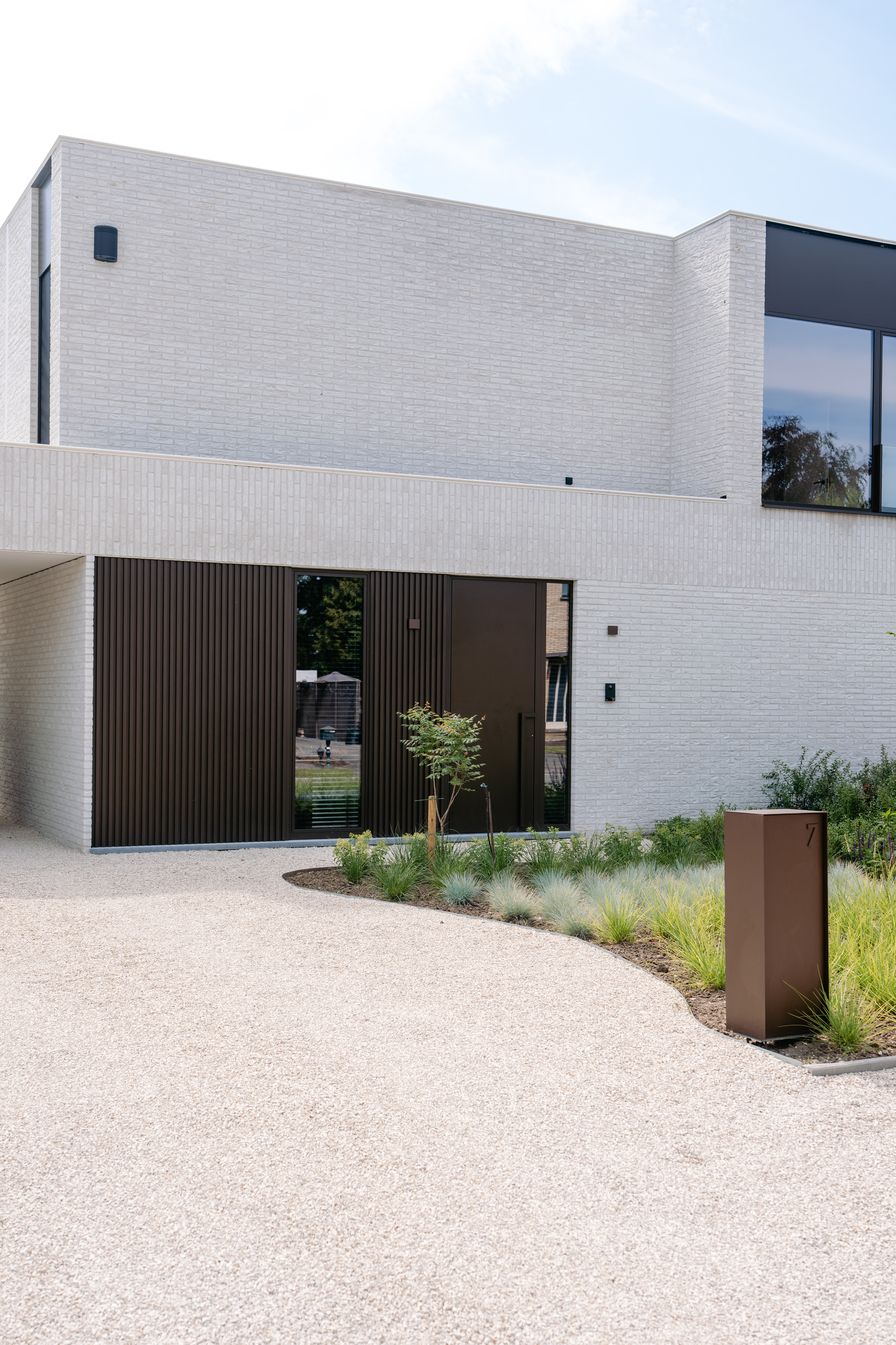 Het moderne huis heeft grote, strakke ramen en een minimalistisch deurontwerp. De ramen zijn ruim en laten veel natuurlijk licht binnen. De deur is geïntegreerd in een verticaal paneelontwerp, wat de eigentijdse uitstraling van het gebouw aanvult.