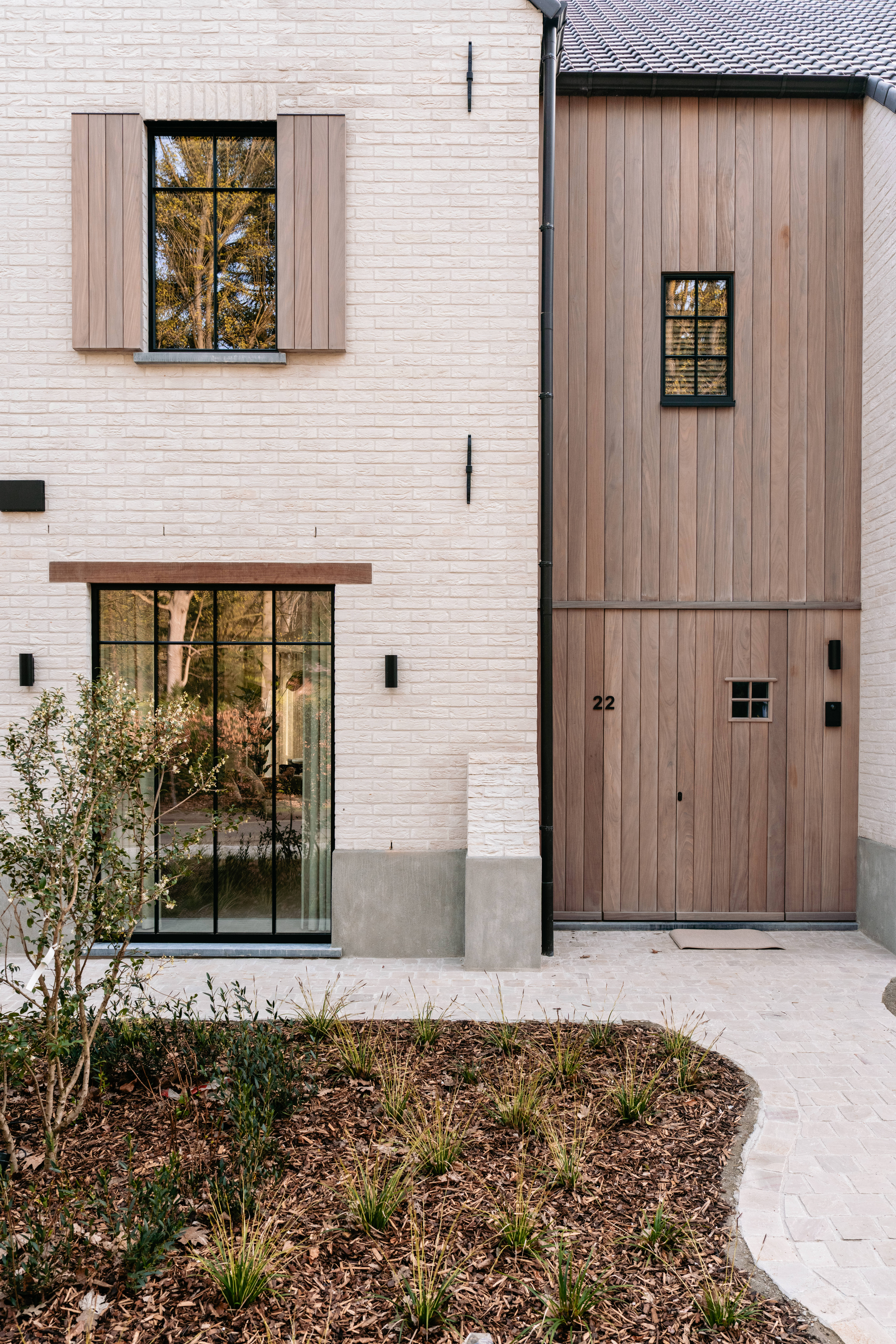 De afbeelding toont een moderne huisgevel met een combinatie van baksteen en houten bekleding. De ramen zijn groot en omlijst met zwart metaal, wat een strakke en eigentijdse uitstraling geeft. De deur is van hout, passend bij de bekleding, en heeft een klein raam. Het geheel is minimalistisch en elegant.