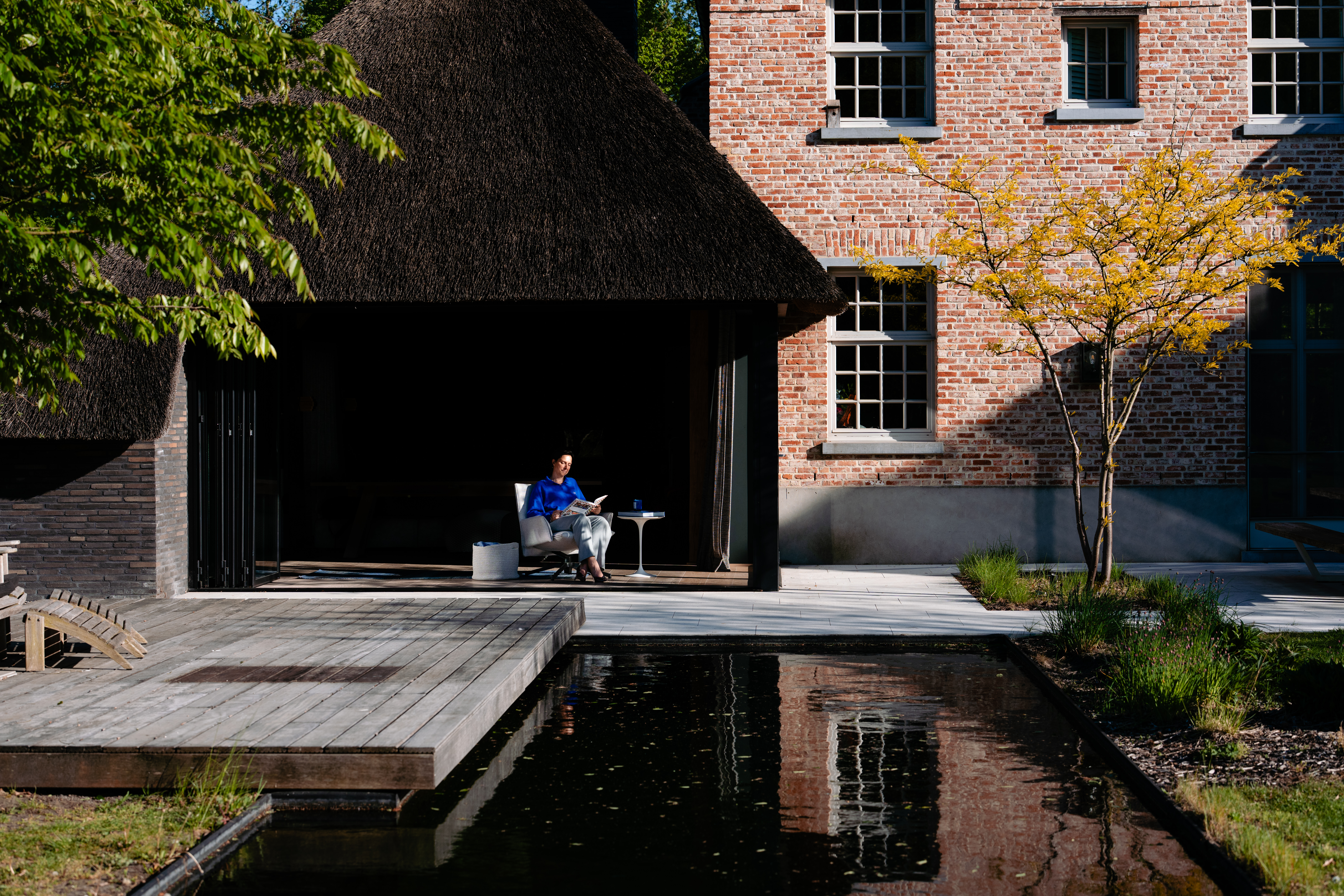 De afbeelding toont een charmant bakstenen huis met meerdere rechthoekige ramen, elk met een klassiek roosterpatroon. De ramen zijn symmetrisch uitgelijnd, wat de traditionele uitstraling van het gebouw versterkt. Naast het huis staat een rieten dakconstructie met grote openstaande deuren, die een naadloze binnen-buitenruimte creëren. De deuren zijn breed en uitnodigend, waardoor natuurlijk licht het interieur binnenstroomt.