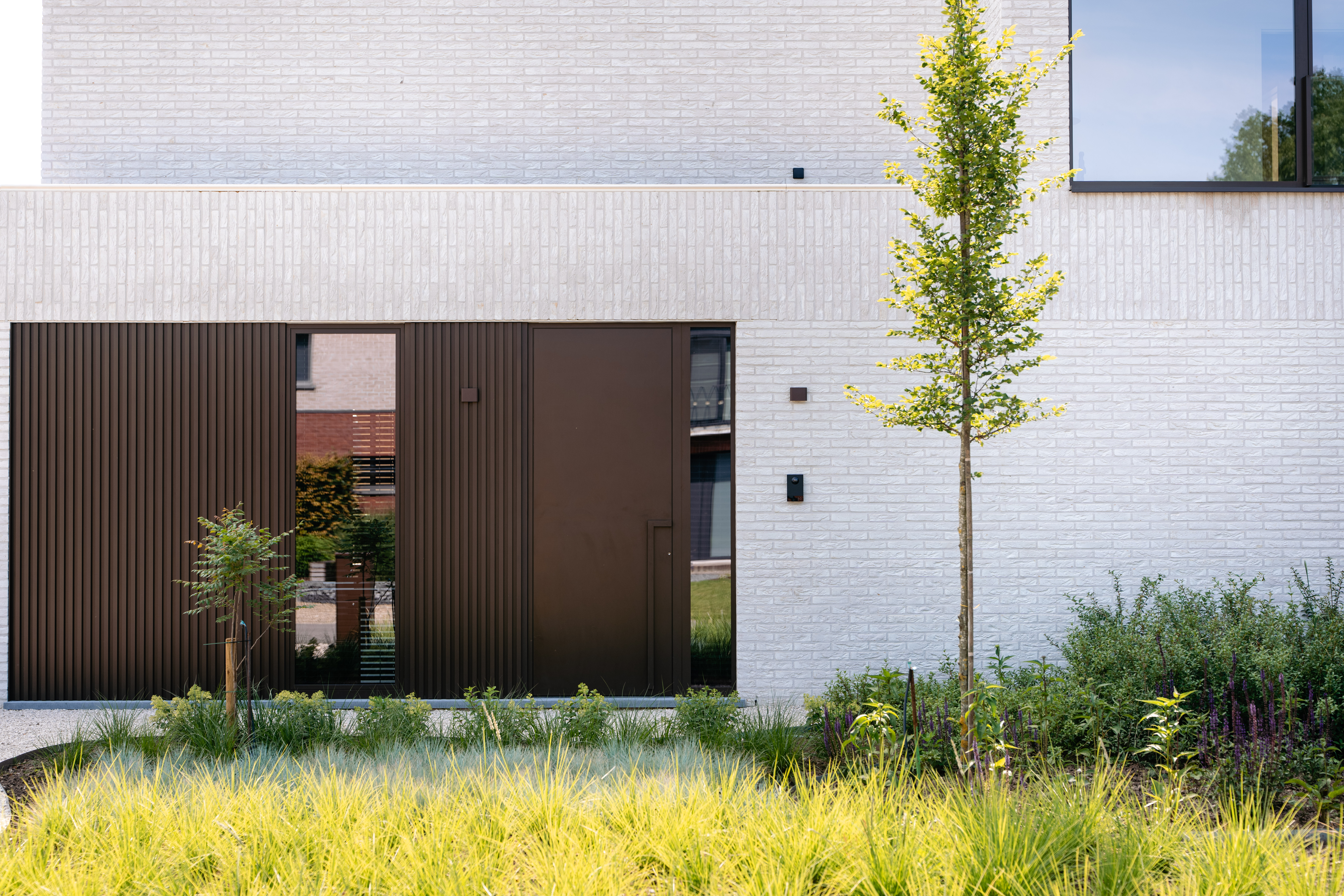 De afbeelding toont een moderne gevel met strakke, donkerbruine deuren en een smal verticaal raam. De deuren zijn geïntegreerd in een witte bakstenen muur, wat een opvallend contrast geeft. Het raam weerspiegelt de omliggende natuur, wat een vleugje natuur toevoegt aan het eigentijdse ontwerp.