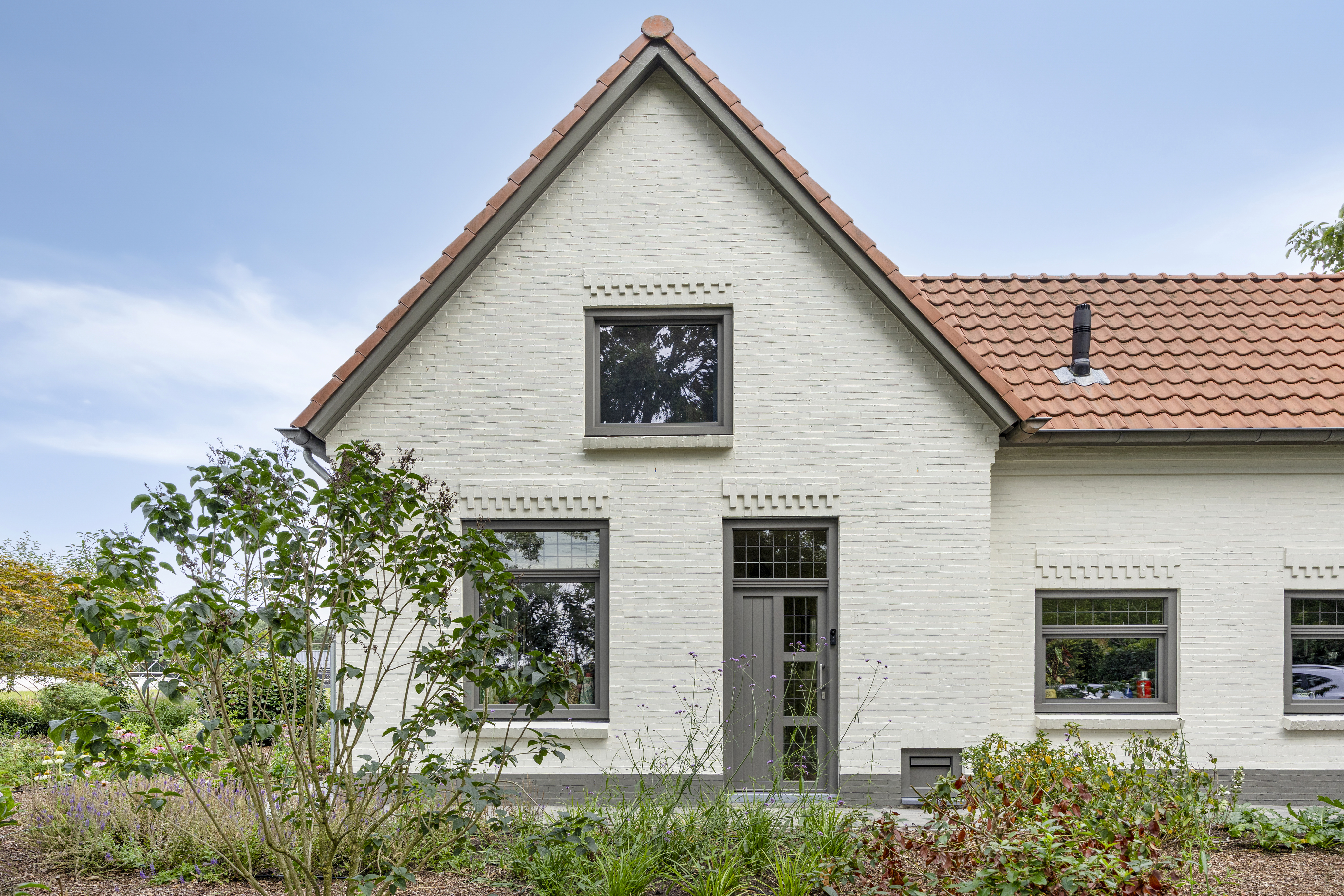 Het huis heeft een klassiek ontwerp met een zadeldak en een witte bakstenen buitenkant. De voorgevel bevat drie kozijnen en een centrale deur. De kozijnen zijn symmetrisch geplaatst, met twee aan weerszijden van de deur en één erboven. De deur is traditioneel met glazen panelen, wat het geheel mooi aanvult.