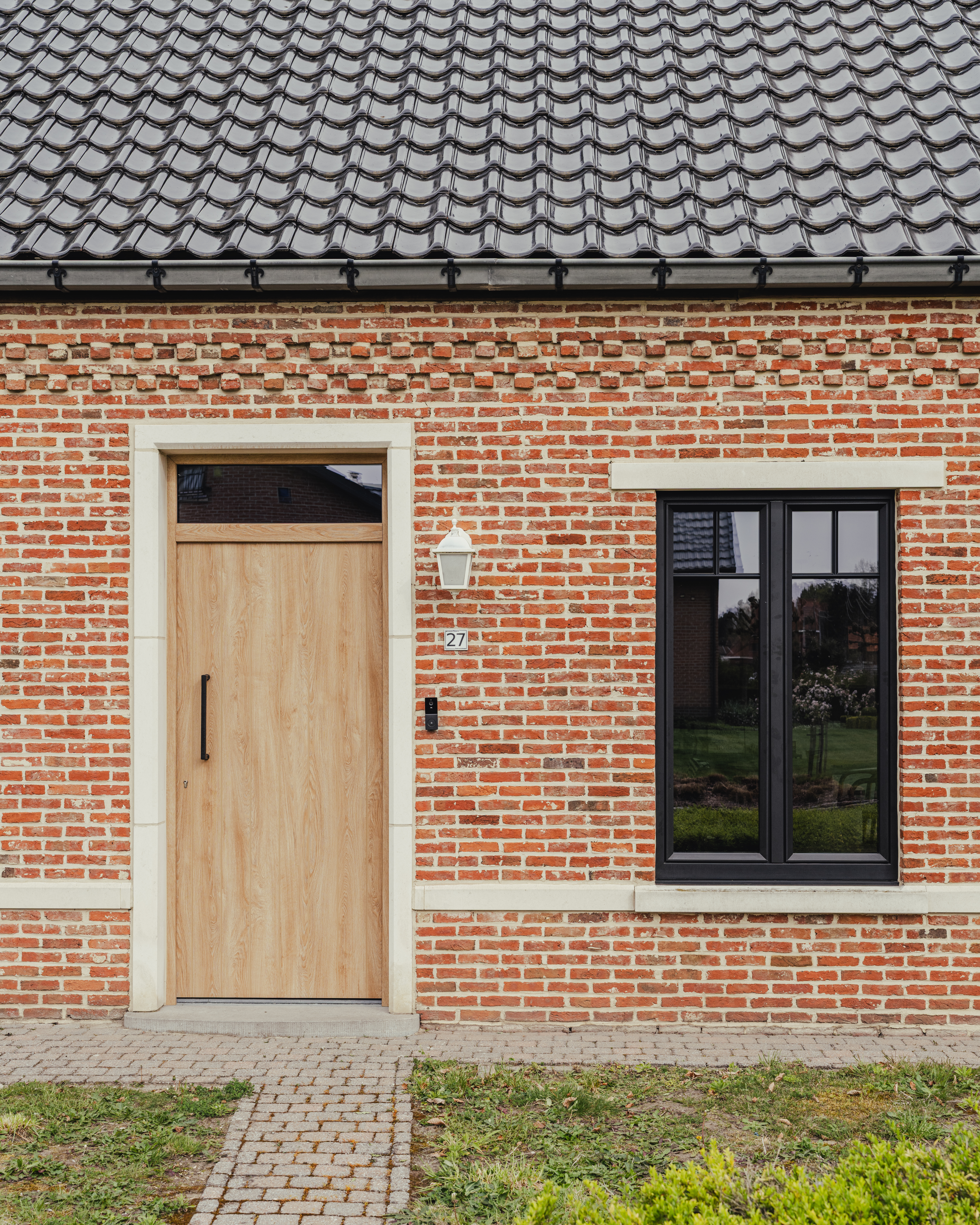 De afbeelding toont een bakstenen gevel van een huis met een moderne houten deur en een zwart omlijst raam. De deur is eenvoudig en elegant, met een verticale handgreep. Het raam ernaast is groot en laat veel licht binnen. De combinatie van rode bakstenen en zwarte omlijstingen geeft een eigentijdse maar klassieke uitstraling.