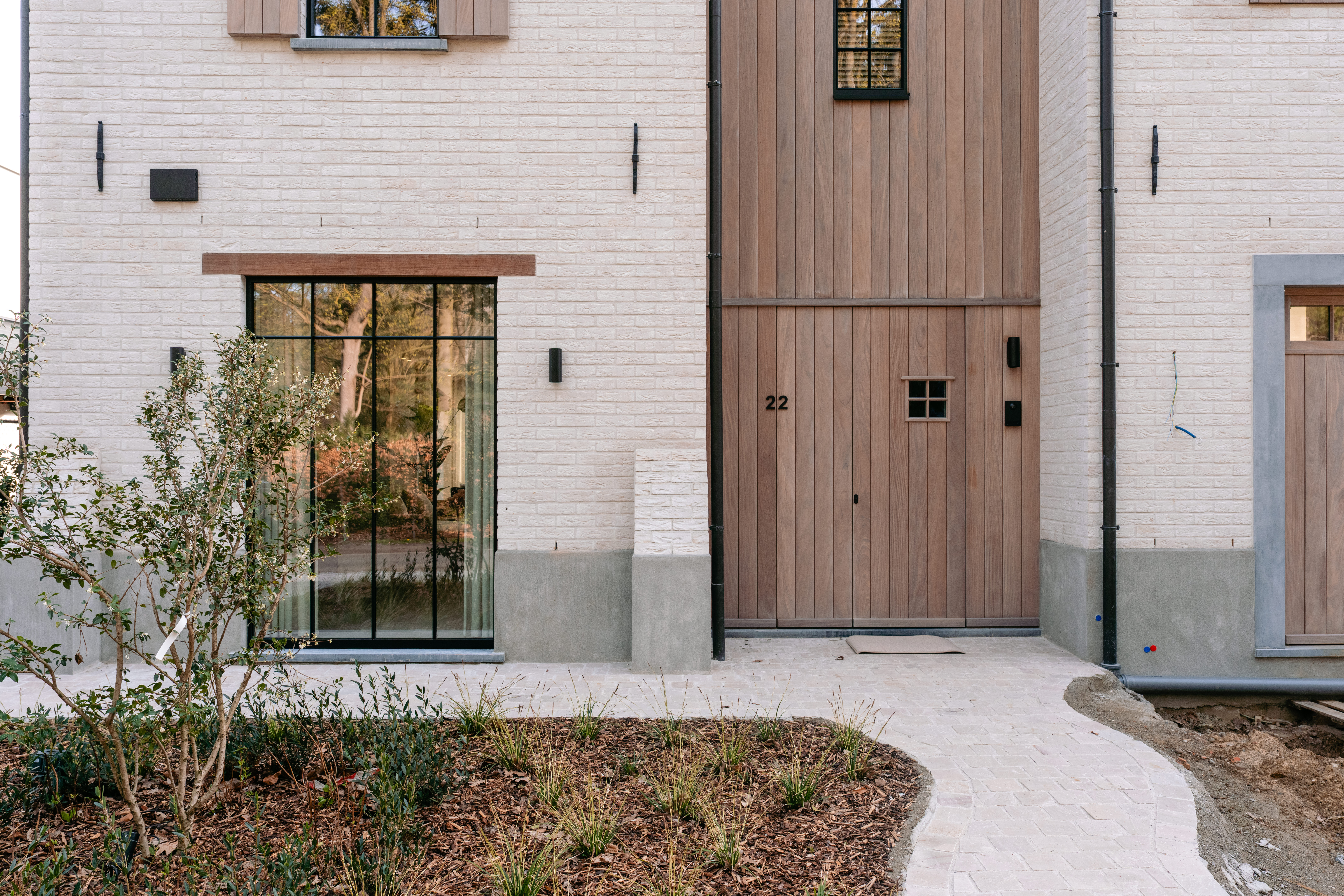 De afbeelding toont een moderne gevel van een huis met een groot raam en een houten deur. Het raam heeft een rasterontwerp dat natuurlijk licht binnenlaat. De houten deur is eenvoudig maar elegant en past goed bij de bakstenen buitenkant. De combinatie van materialen geeft het huis een eigentijdse en uitnodigende uitstraling.