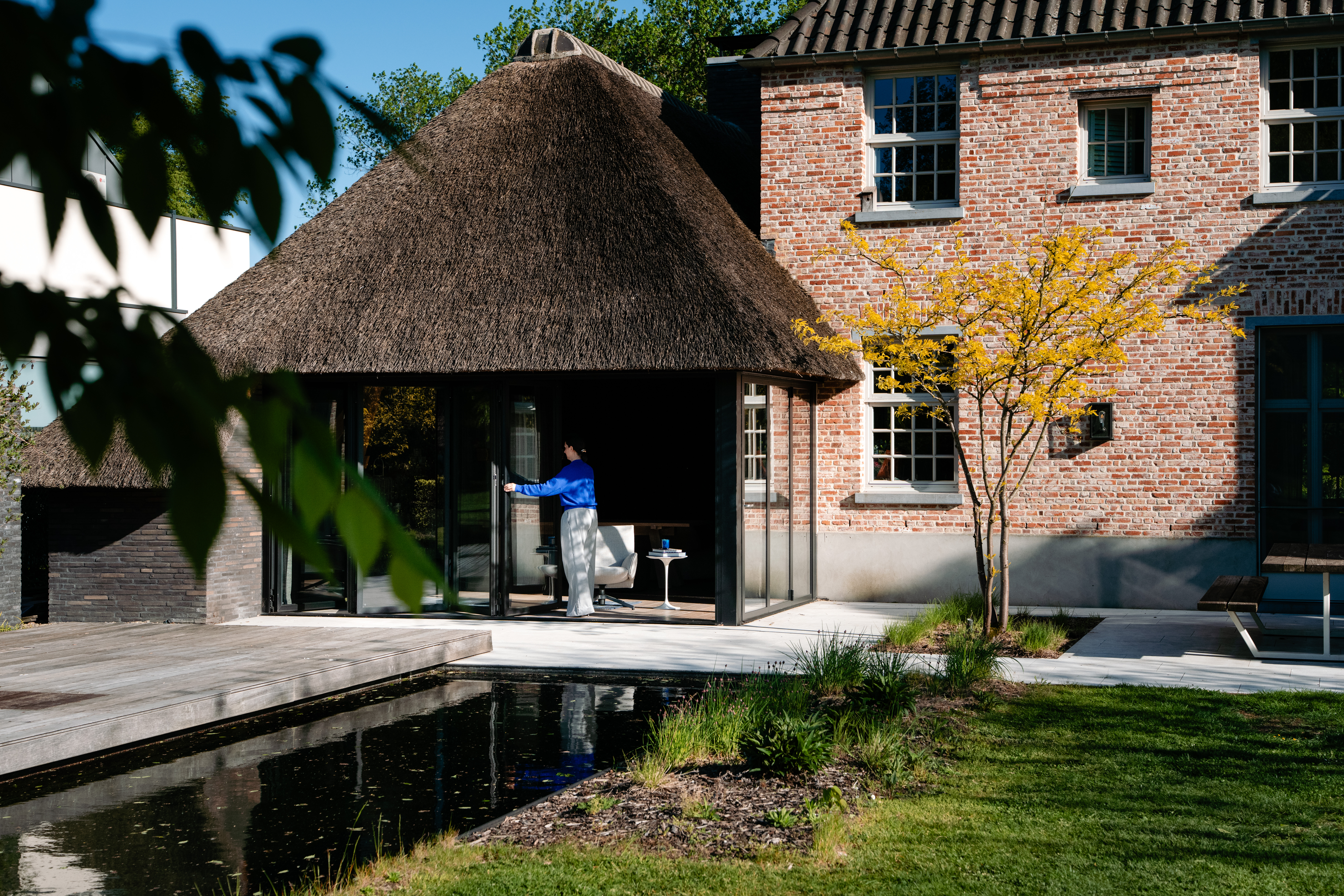 De afbeelding toont een charmant bakstenen huis met een rieten dakuitbreiding. Het huis heeft meerdere witomrande ramen en een groot schuifraam dat naar een terras leidt. De ramen zijn symmetrisch geplaatst, wat de rustieke maar elegante uitstraling van het gebouw versterkt. Het schuifraam opent naar een serene tuin met een vijver, waardoor binnen- en buitenruimtes naadloos in elkaar overgaan.