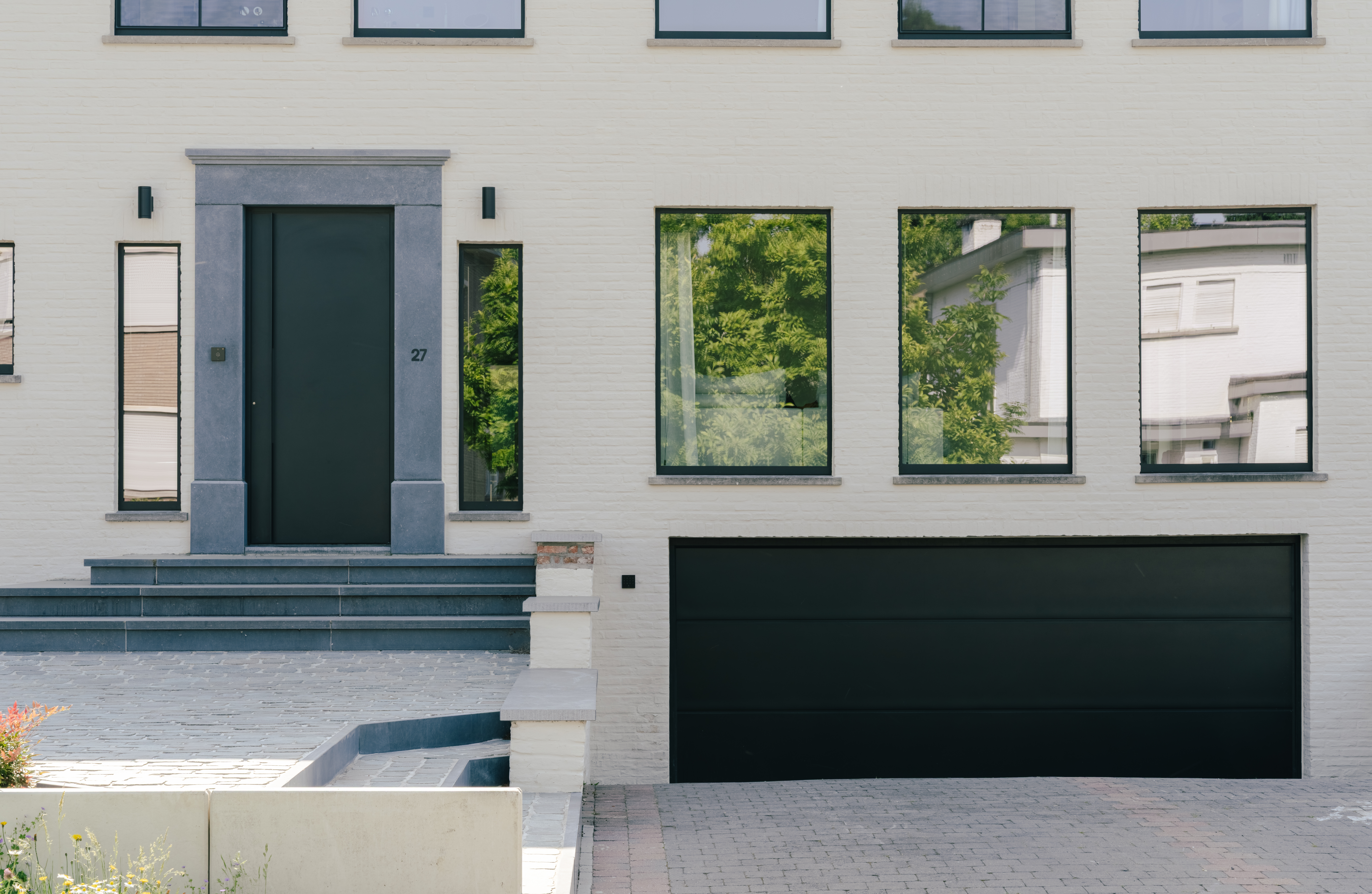 De afbeelding toont een moderne gevel van een huis met grote, rechthoekige ramen en een strakke zwarte deur. De ramen zijn symmetrisch geplaatst, waardoor er veel natuurlijk licht binnenkomt. De deur is centraal geplaatst, geflankeerd door twee smalle ramen, wat het eigentijdse ontwerp versterkt. De garagepoort eronder heeft dezelfde kleur als de deur, wat zorgt voor een samenhangend geheel.