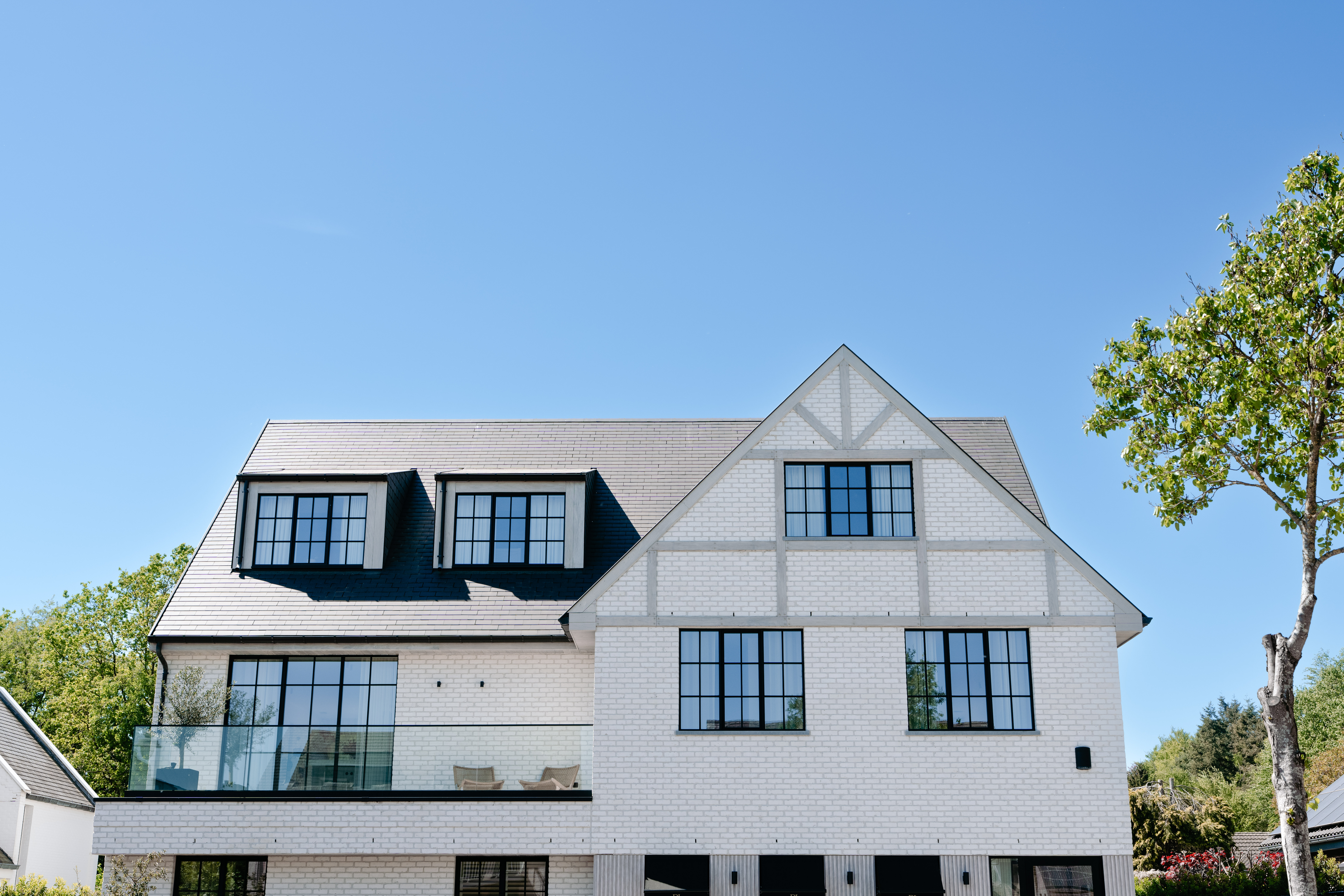 Het huis heeft grote, zwartomrande ramen die een moderne toets geven aan de traditionele architectuur. De ramen zijn symmetrisch geplaatst, wat de esthetische aantrekkingskracht vergroot. De voordeur is niet zichtbaar, maar de ramen domineren de gevel en laten veel natuurlijk licht binnen.