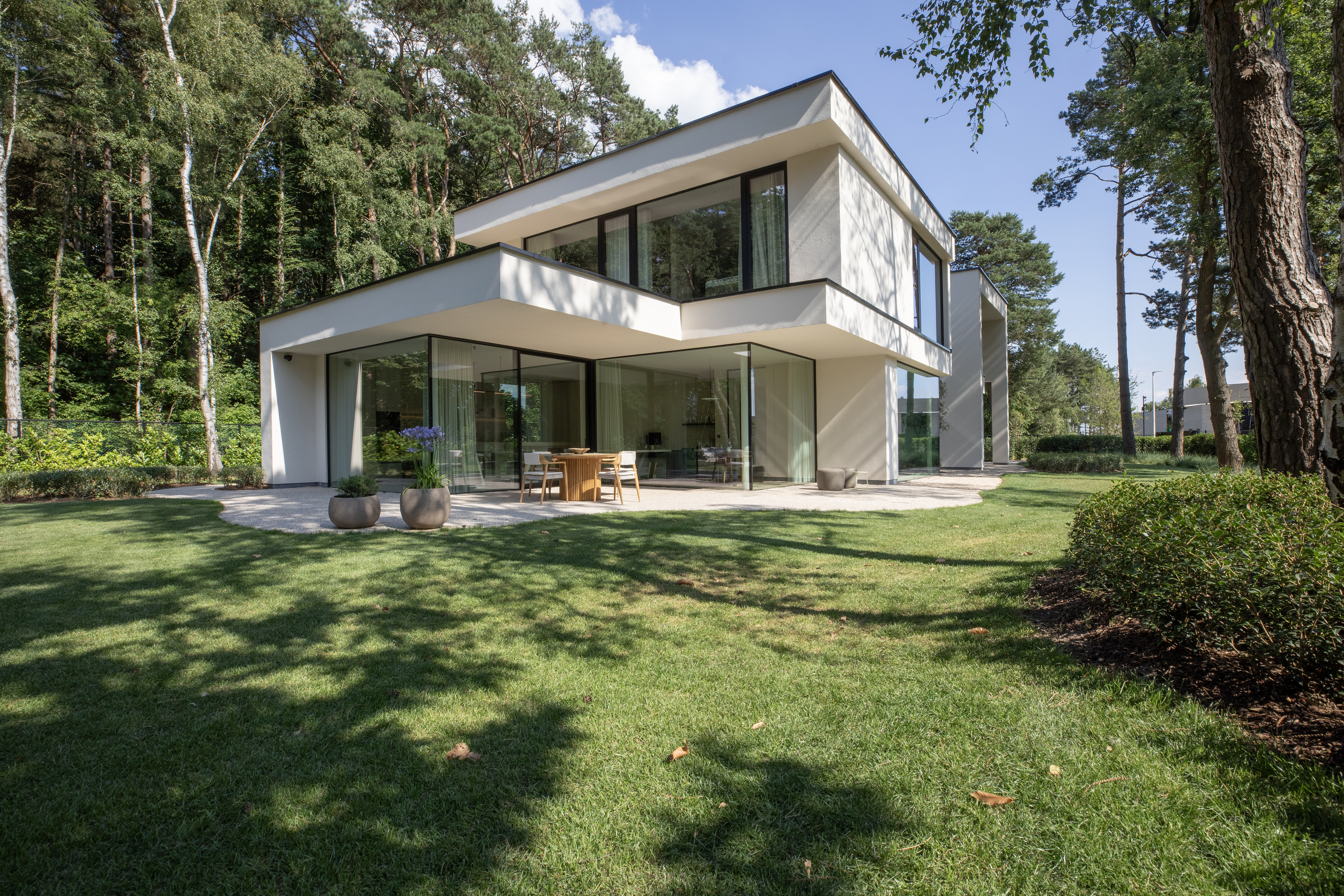 Het moderne huis heeft grote glazen schuiframen en deuren, die een naadloze verbinding tussen binnen en buiten creëren. De ramen zijn ruim en laten veel natuurlijk licht binnen. Het strakke ontwerp past bij de minimalistische architectuur, met de glazen elementen die een helder zicht op het omliggende groen bieden.